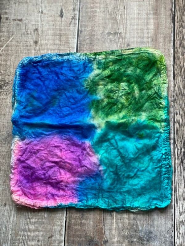 a stack of hand dyed Mawata silk hankies, in green, purple and two tones of blue, on a table