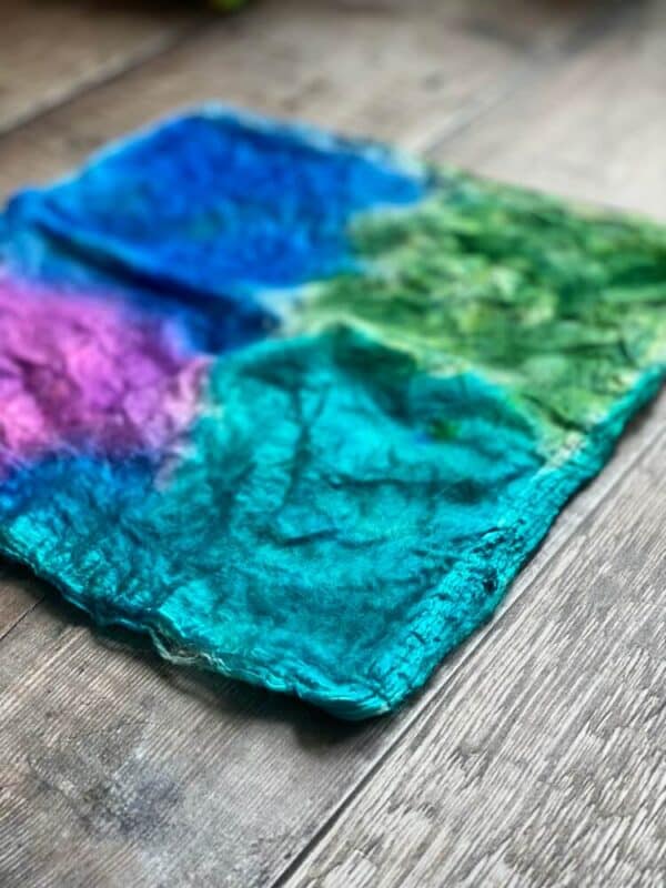 closeup of a hand dyed mawata silk hankie in blues, green and purple. The light blue corner is closest to the camera.