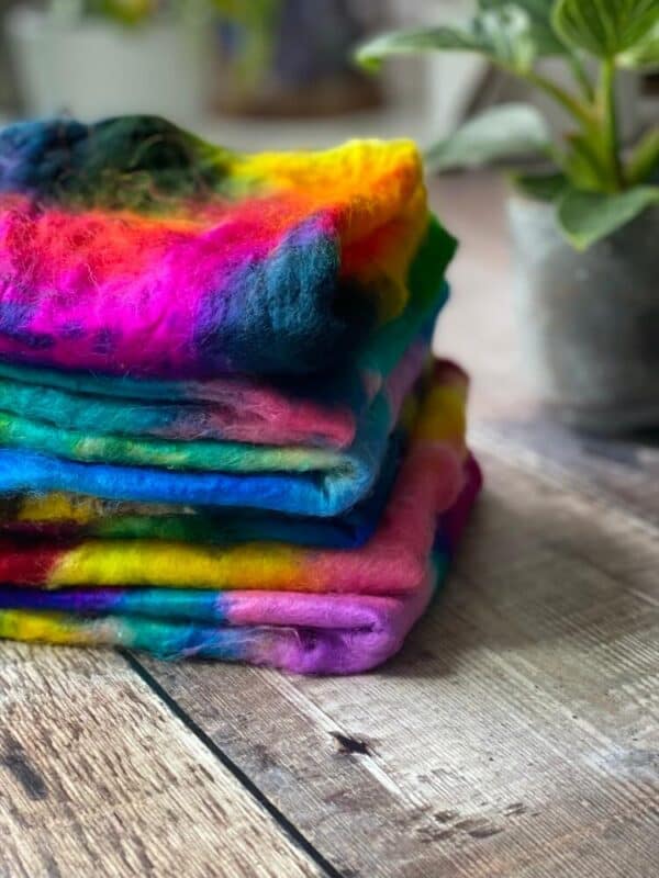 a stack of different hand dyed Mawata silk hankies on a table, with a plant in the background of Eleanor Shadow's studio